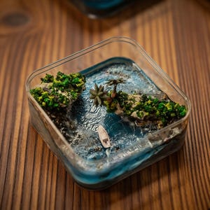 Puede incluir: Diorama oceánico en miniatura en un recipiente cuadrado transparente. La escena incluye agua azul, pequeñas islas con follaje verde, una palmera y un pequeño bote blanco. El diorama está sobre una superficie de madera.