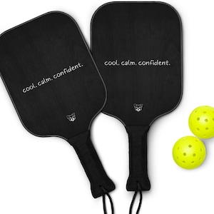 May include: Two black pickleball paddles with the text "cool. calm. confident." and a small logo. Two yellow pickleballs are also in the image. The paddles have black handles with a black cord attached.