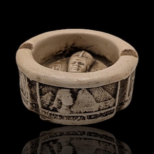 May include: A cream-coloured ashtray with a detailed relief of a figure inside the bowl. The exterior features an etched design of pyramids and hieroglyphs. The ashtray is round with a small notch for resting a cigarette.