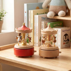 May include: Two wooden carousel music boxes featuring small hand-painted figures. One carousel has a red base and roof, while the other is natural light wood. Both have a central pillar with rotating horses and figures, and a small winding knob.
