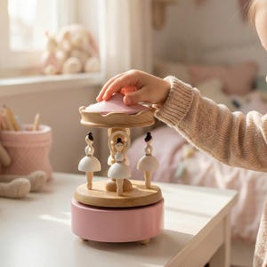 May include: A pink and wood-toned carousel music box with three ballerina figures. The carousel has a pink base, a wooden midsection, and a pink top. The ballerina figures are white with black hair. The carousel is on a white table.
