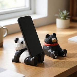 May include: Two carved wooden phone holders. One is a panda bear with black and white coloring, holding a black phone. The other is a black bear with a red collar. Both are sitting on a wooden surface.
