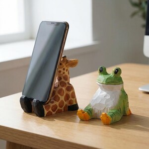 May include: A wooden giraffe-shaped phone stand with a smartphone in place, next to a carved green frog figurine. The giraffe stand has brown spots and black feet. The frog is green and white with orange feet.