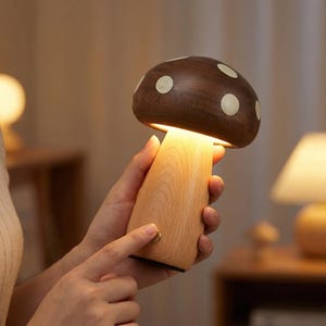 Puede incluir: Una lámpara con forma de hongo con base de madera y una tapa marrón oscuro con lunares blancos. La lámpara está encendida, emitiendo un brillo cálido y suave. La lámpara se sostiene en una mano.