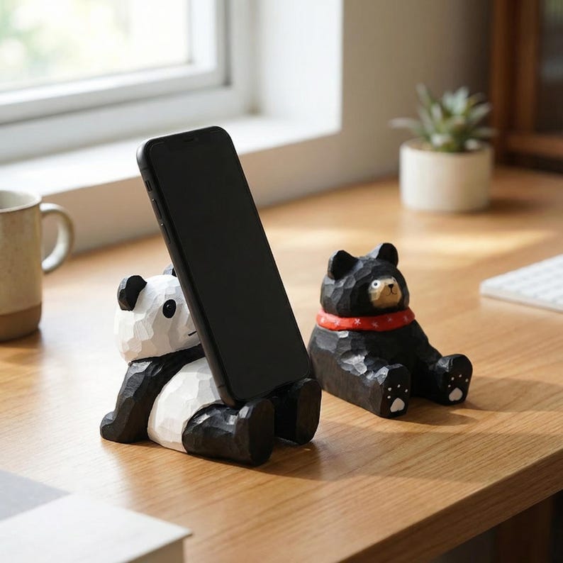 May include: Two carved wooden phone holders. One is a panda bear with black and white coloring, holding a black phone. The other is a black bear with a red collar. Both are sitting on a wooden surface.