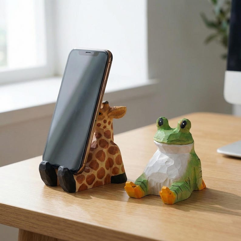 May include: A wooden giraffe-shaped phone stand with a smartphone in place, next to a carved green frog figurine. The giraffe stand has brown spots and black feet. The frog is green and white with orange feet.