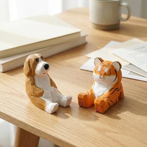 May include: Two wooden animal figurines, a dog and a tiger, sit on a light-colored wooden surface. The dog is brown and white, while the tiger is orange and white with black stripes. Both are carved in a seated position.
