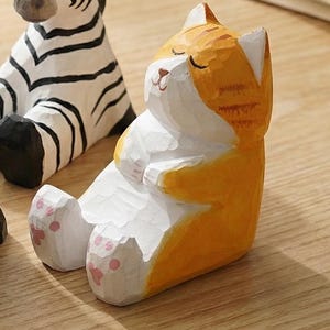 May include: A hand-carved wooden cat figurine in a relaxed pose, painted in orange and white. The cat is sitting with its eyes closed and paws together. The figurine has pink paw pads. A zebra figurine is partially visible in the background.