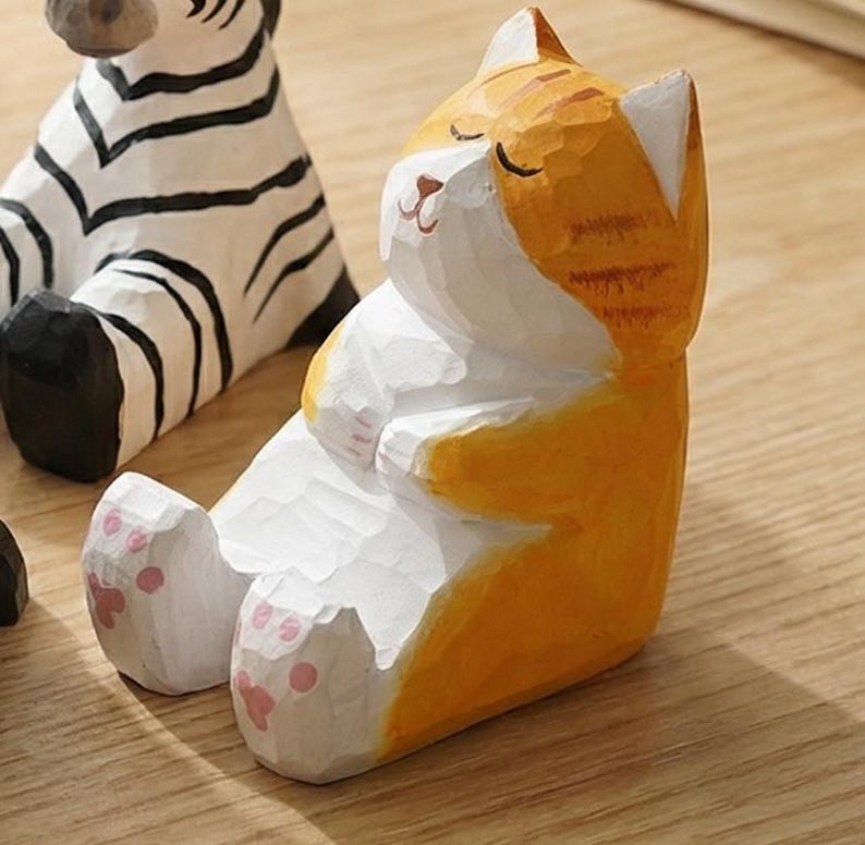 May include: A hand-carved wooden cat figurine in a relaxed pose, painted in orange and white. The cat is sitting with its eyes closed and paws together. The figurine has pink paw pads. A zebra figurine is partially visible in the background.