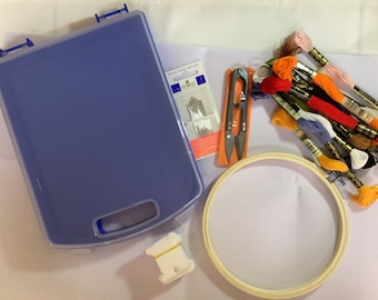 May include: A blue plastic storage container, a wooden embroidery hoop, a pack of embroidery needles, a pair of small scissors, and a variety of colorful embroidery floss.