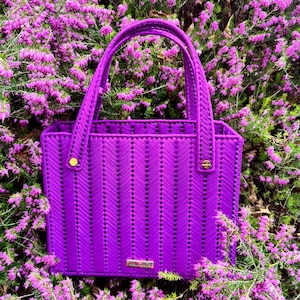 Puede incluir: Un bolso morado vibrante con un diseño tejido y asas a juego. El bolso presenta herrajes dorados y una pequeña etiqueta rectangular. El bolso está sobre un fondo de flores moradas y follaje verde.