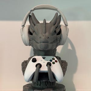 May include: A gray dragon-themed headset and controller stand. The stand holds a white Xbox controller and a pair of white headphones. The base of the stand has engraved text. The dragon's head is the focal point.