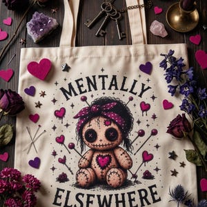 Könnte beinhalten: Eine beigefarbene Stofftasche mit einer aufgedruckten Illustration einer Voodoo-Puppe mit Knopfaugen, einem Herz auf der Brust und einem gepunkteten Stirnband. Der Text "MENTALLY ELSEWHERE" ist in Schwarz über und unter der Puppe aufgedruckt.