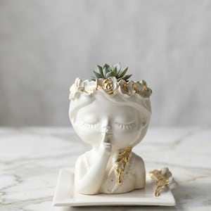 Può includere: Vaso in ceramica bianca a forma di volto femminile, con occhi chiusi e un dito sulle labbra. Il vaso presenta una corona floreale con dettagli dorati e contiene una piccola pianta succulenta. Poggia su una base quadrata bianca.