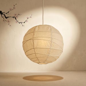 May include: A spherical cream paper lantern suspended from a white cable. The lantern emits a warm light, and has horizontal bands. The shadow of the lantern is visible on the floor. The background features a painted floral design.
