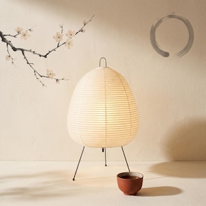 Könnte beinhalten: Eine cremefarbene Papierlaterne mit einem schwarzen Metall-Dreibein-Sockel. Die Laterne hat eine abgerundete, eiförmige Form und ist von innen beleuchtet. Eine kleine, braune Keramikschale steht in der Nähe. Der Hintergrund zeigt einen Zweig mit weißen Blüten.