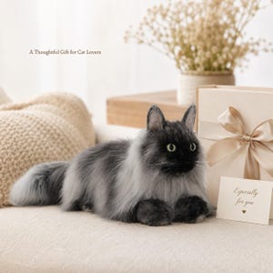 May include: A plush, gray and black cat with green eyes, lying on a cream-colored surface. The cat has long fur and a fluffy tail. A gift box with a bow and a card that says "Especially for you" are next to the cat. The text "A Thoughtful Gift for Cat Lovers" is above the cat.