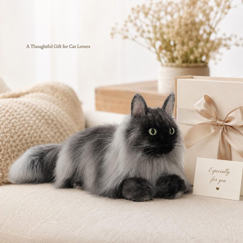 May include: A plush, gray and black cat with green eyes, lying on a cream-colored surface. The cat has long fur and a fluffy tail. A gift box with a bow and a card that says "Especially for you" are next to the cat. The text "A Thoughtful Gift for Cat Lovers" is above the cat.