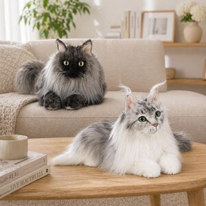 May include: Two plush cat toys. One is a gray and black cat with a fluffy coat, sitting on a beige sofa. The other is a gray and white cat with green eyes, lying on a wooden table. The cats have realistic features.