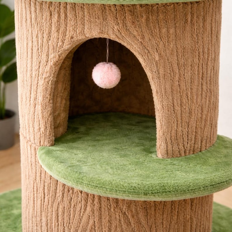 May include: A brown cat tree with a tree trunk design, featuring a green carpeted base and a cozy interior. A pink pom-pom hangs from the top, providing playful interaction. The tree has a textured, wood-like appearance.