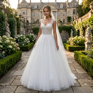 Peut inclure: Une robe de mariée blanche avec un corsage encolure en V, des manches courtes et une jupe en tulle ample. La robe présente des détails de dentelle sur le corsage et la jupe. Un voile transparent est attaché aux cheveux de la mariée. En arrière-plan, un chemin pavé et un grand bâtiment.