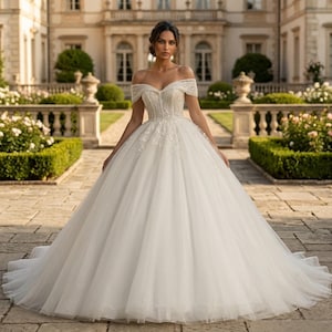 May include: A white off-the-shoulder wedding gown with a beaded bodice and a full, flowing skirt. The dress features delicate embellishments and a long train. The bride is standing in a garden setting with a large building in the background.