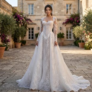 May include: A white lace wedding dress with long sleeves and a sweetheart neckline. The dress features a fitted bodice with button details and a flowing skirt. The bride is standing in a courtyard with stone pavers and a building in the background.