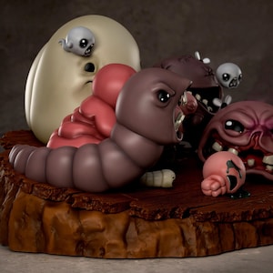 May include: A detailed figurine set featuring several stylized, cartoonish figures. The set includes a large, off-white egg-shaped character, a pink, fleshy mass, a large brown worm, and several smaller, grotesque figures. The base is a brown, textured platform.