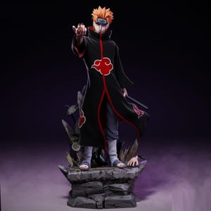 May include: A detailed figurine of a character from the Naruto anime series. The figure is posed on a rocky base, wearing a black cloak with red accents and cloud symbols. The character has spiky orange hair and is holding a weapon.