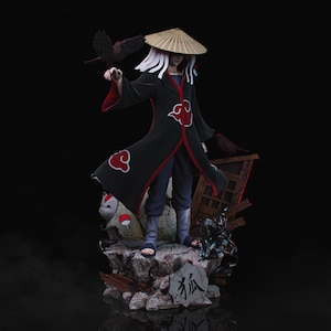 Puede incluir: Figura detallada de un personaje con una túnica negra con detalles rojos, un sombrero de paja y cabello blanco. La figura sostiene un pájaro negro y se encuentra sobre una base rocosa con un símbolo de zorro. El fondo es negro.