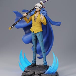 Può includere: Statuetta dettagliata di un personaggio della serie anime One Piece. La figura indossa una giacca gialla, jeans blu e un cappello bianco con pois neri. Tiene in mano un bastone nero con stelle bianche e una spada viola. La figura è su una base rocciosa con accenti di fiamma blu.