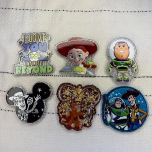 May include: A selection of six colourful, glittery pins featuring characters and phrases from the Toy Story franchise. The pins include images of Jessie, Buzz Lightyear, Woody, and Mickey Mouse, along with the phrase "I Love You to Infinity and Beyond."