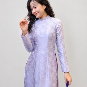 Può includere: Un ao dai color lavanda a maniche lunghe con un sottile motivo floreale jacquard bianco, un colletto alla coreana impreziosito da piccole perline bianche e profondi spacchi laterali che rivelano uno strato di tessuto viola scuro.