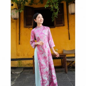 May include: A long-sleeved, floor-length tunic dress featuring a vibrant pink base with a repeating silver floral pattern, paired with light blue trousers. The garment has a high collar and a side slit that reveals the light blue pants underneath.