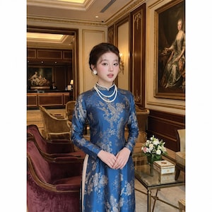 May include: A long-sleeved blue dress featuring a high collar and a repeating floral pattern in a metallic gold tone. The garment is styled with a double-strand white pearl necklace and small white flower-shaped stud earrings.