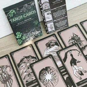 May include: A deck of "TAROT CARDS" with a black and green box, inspired by the video game "Phasmophobia." The cards feature black and white illustrations on a pink background, with titles like "THE FOOL" and "THE HANGED MAN."