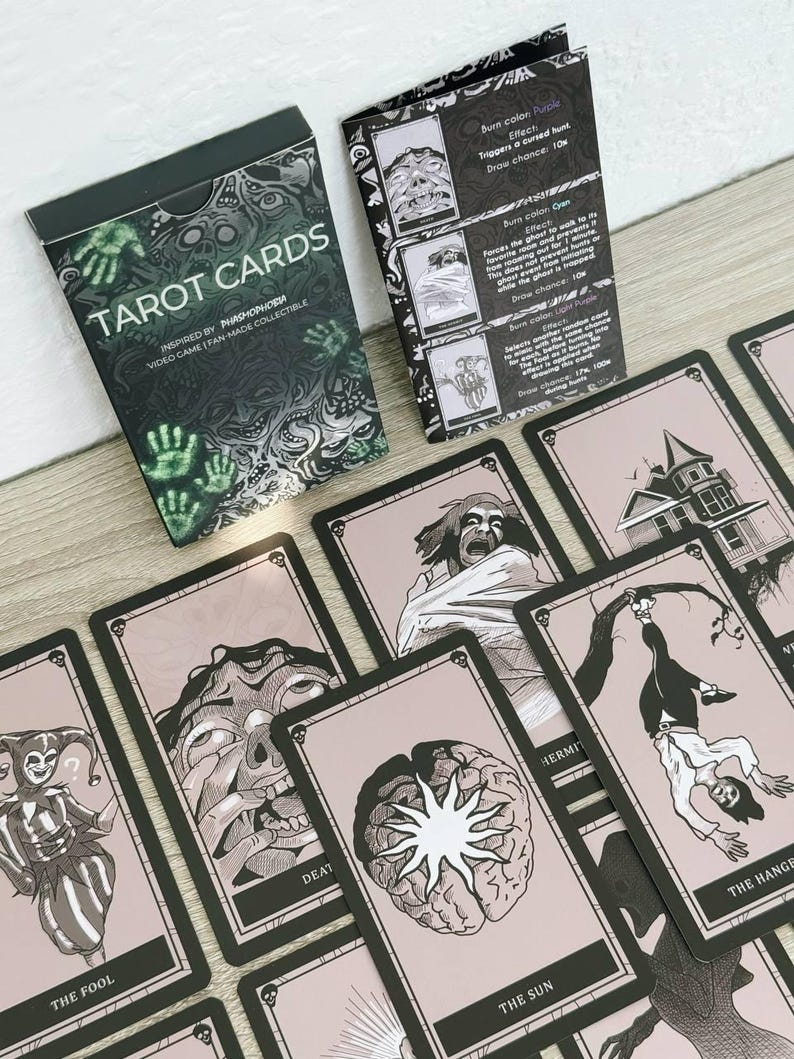 May include: A deck of "TAROT CARDS" with a black and green box, inspired by the video game "Phasmophobia." The cards feature black and white illustrations on a pink background, with titles like "THE FOOL" and "THE HANGED MAN."