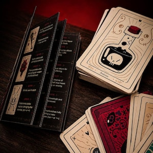 May include: A deck of illustrated game cards featuring a bottle labeled Ambrosia with a skull icon, alongside a black instruction booklet with white text and red accents. The cards display various graphic designs including a red card with an eye.