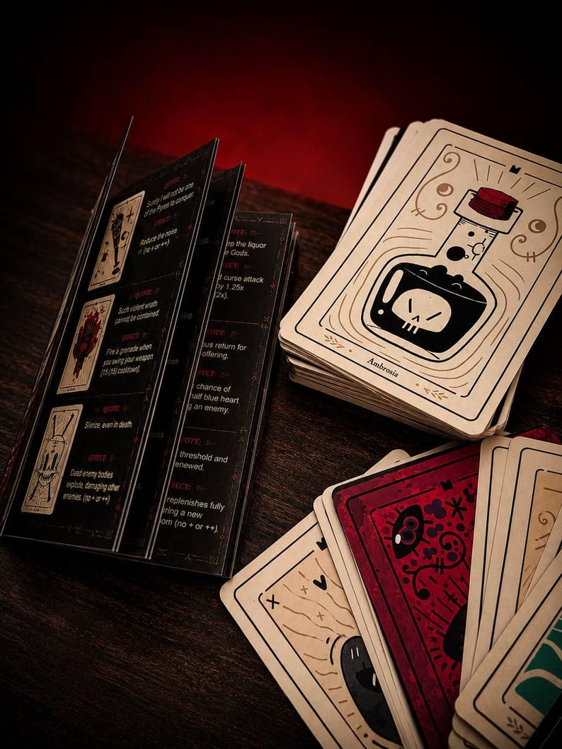 May include: A deck of illustrated game cards featuring a bottle labeled Ambrosia with a skull icon, alongside a black instruction booklet with white text and red accents. The cards display various graphic designs including a red card with an eye.