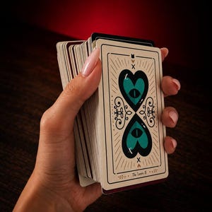 May include: A deck of tarot cards with a cream-colored back featuring a symmetrical design of two teal hearts, each containing a dark eye. The card at the front displays the text The Lovers II, surrounded by black line art and small heart icons.