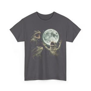 May include: Dark gray t-shirt featuring a whimsical design of three frogs with the moon in the background. The frogs appear to be singing or celebrating, with one frog raising its arms towards the moon. The design is detailed and artistic.