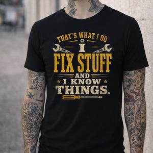 Könnte beinhalten: Schwarzes T-Shirt mit dem Text "THAT'S WHAT I DO I FIX STUFF AND I KNOW THINGS." Der Text ist in Gelb und Weiß gehalten, mit Abbildungen eines Schraubenschlüssels und eines Schraubendrehers. Das T-Shirt ist kurzärmelig.