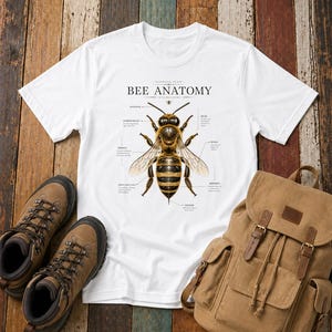 Puede incluir: Una camiseta blanca de manga corta con una ilustración científica detallada de una abeja en el centro. El gráfico incluye el texto "BEE ANATOMY" y "APIS MELLIFERA" con varias etiquetas que señalan partes específicas del cuerpo de la abeja.
