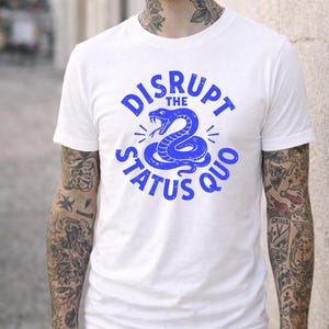 May include: White t-shirt featuring a blue snake graphic and the text "DISRUPT THE STATUS QUO". The design has a distressed effect. The t-shirt is a crew neck style, a popular casual wear choice.