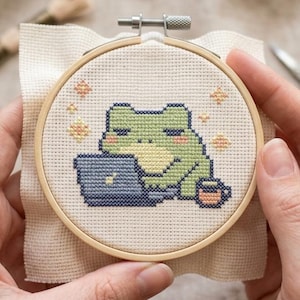 Puede incluir: Un bastidor de bordado de punto de cruz con una rana verde trabajando en un portátil, con una taza de café al lado. La rana está bordada en tela color crema, colocada en un bastidor de madera. El diseño incluye pequeños acentos de estrellas amarillas y naranjas.
