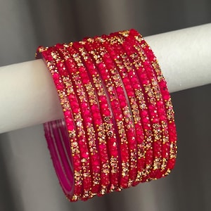 May include: A set of eight vibrant fuchsia pink bangles, each featuring a textured surface adorned with small, sparkling gold-toned accents arranged in a repeating pattern around the circumference of every individual circular bracelet.