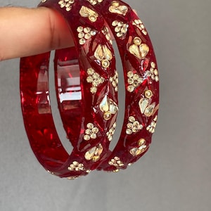May include: Two translucent deep red bangles featuring a faceted surface texture. Each bangle is decorated with embedded clear, sparkling crystals arranged in geometric patterns across the outer circumference of the circular jewellery pieces.