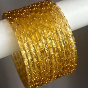 May include: A set of seven translucent yellow bangles featuring a repeating diamond-shaped textured pattern across the surface. Each bangle is accented with small, raised gold-coloured dots arranged in vertical lines between the diamond shapes.