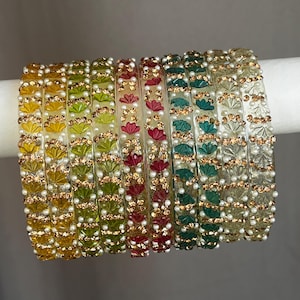 May include: A set of ten translucent bangles featuring a repeating fan-shaped pattern in yellow, green, red, and clear tones. Each bangle is accented with small, round, white faux pearls and tiny, sparkling gold-toned beads in a linear arrangement.