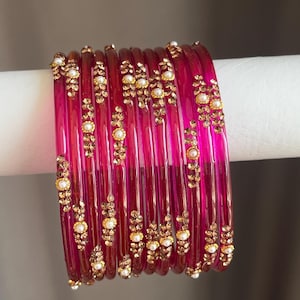 May include: A set of ten translucent fuchsia pink bangles, each featuring a vertical line of small gold-toned metallic embellishments interspersed with tiny white faux pearls, creating a uniform decorative pattern across the circular bracelets.